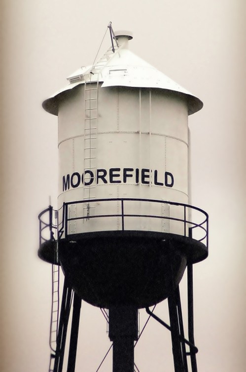 A tall water tower with the word "MOOREFIELD" painted on its side, set against a gray sky.