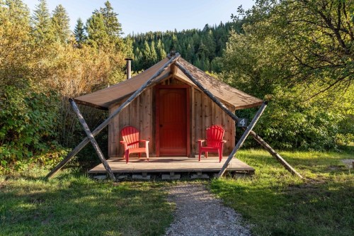 A cozy wooden cabin with a red door and two red chairs on the porch, surrounded by trees and greenery.