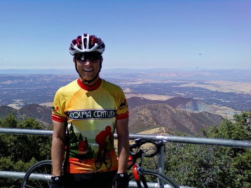 A smiling cyclist in a colorful jersey stands near a bike, overlooking a scenic mountain view.