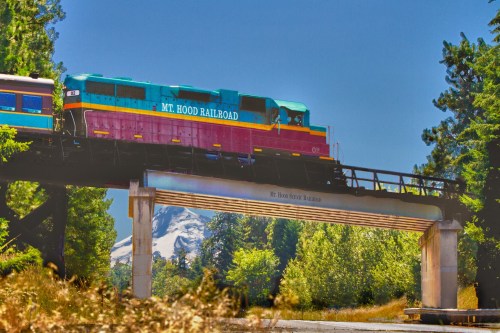 A colorful train labeled "Mt. Hood Railroad" crosses a bridge with a mountain in the background and lush greenery around.