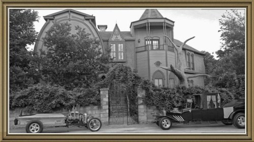 A vintage house with unique architecture, flanked by two classic cars, surrounded by lush greenery.