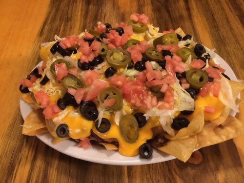 A plate of nachos topped with melted cheese, jalapeños, black olives, lettuce, and diced tomatoes on a wooden table.