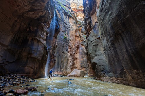 A person stands in a narrow canyon with towering walls, sunlight streaming down into the water below.