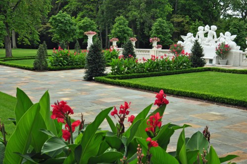 A lush garden featuring vibrant red flowers, neatly trimmed hedges, and elegant white sculptures amidst greenery.