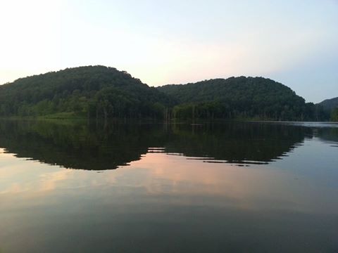 Serene lake reflecting green hills under a soft pastel sky at dusk.