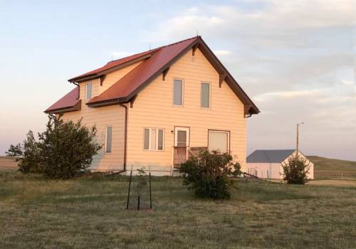 A two-story house with a red roof, surrounded by grass and small shrubs, set against a clear sky.