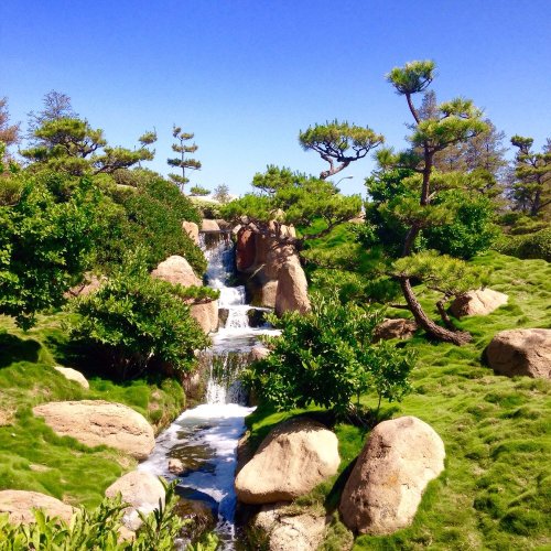 A serene landscape featuring a small waterfall, lush greenery, and rocky formations under a clear blue sky.