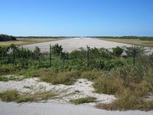 A clear view of an empty runway surrounded by grass and a chain-link fence under a blue sky.