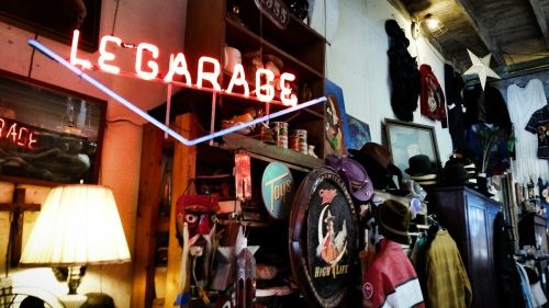 A vintage shop interior featuring a neon "Le Garage" sign, hats, and various eclectic items on display.