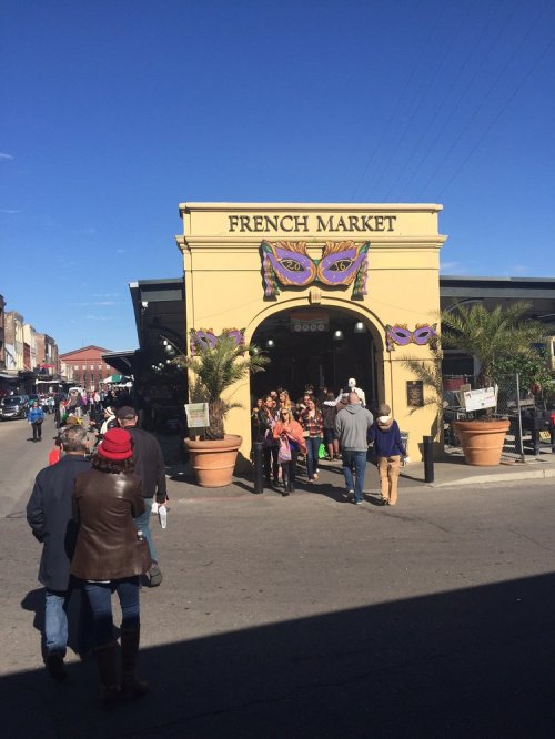 Entrance to the French Market, bustling with visitors, adorned with colorful masks and palm trees under a clear blue sky.