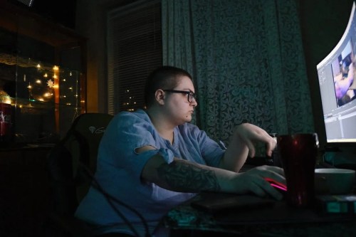 A person with short hair and glasses works intently at a computer in a dimly lit room, surrounded by soft lights.