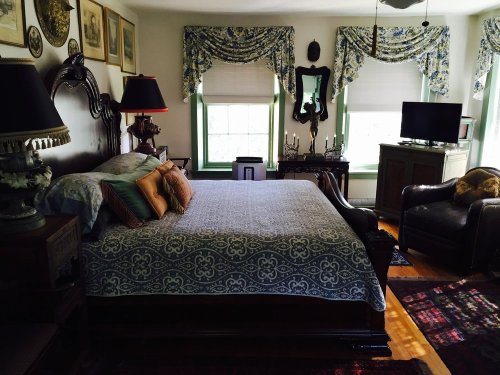 Cozy bedroom with a large bed, decorative pillows, vintage decor, and a TV, featuring green window frames and patterned curtains.