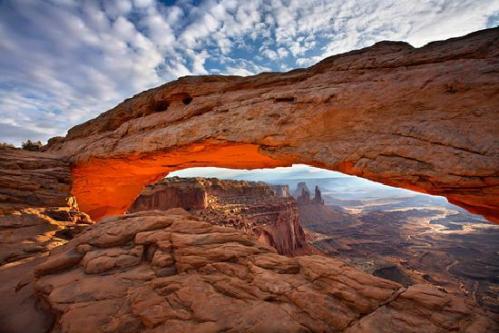 A stunning rock arch illuminated by sunset, overlooking a vast desert landscape with distant mesas and dramatic clouds.