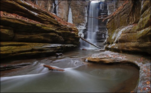 A serene waterfall cascades down rocky cliffs into a calm pool, surrounded by autumn leaves and smooth stones.