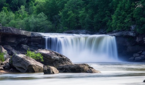 A serene waterfall cascades over rocks, surrounded by lush green trees and a calm river.