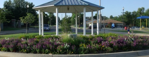 A gazebo surrounded by colorful flower beds in a park, with trees and a street in the background.
