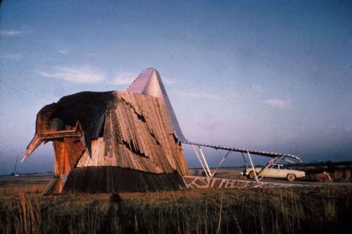 A quirky, bird-shaped building with a sloped roof, set in a grassy landscape beside a vintage car.