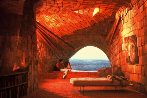 A unique, warm-toned interior with textured walls, a bed, and a person gazing out a large arched window at a landscape.