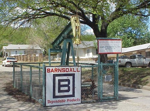 A green oil pump jack behind a fence, with a sign reading "Barnsdall Dependable Products" and a nearby informational sign.