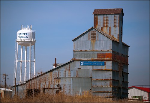 Rusty grain elevator next to a water tower, set against a clear blue sky in a rural landscape.