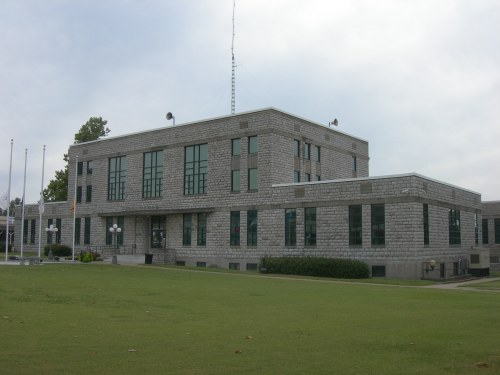 A modern stone building with large windows, surrounded by green grass and flagpoles, under a cloudy sky.