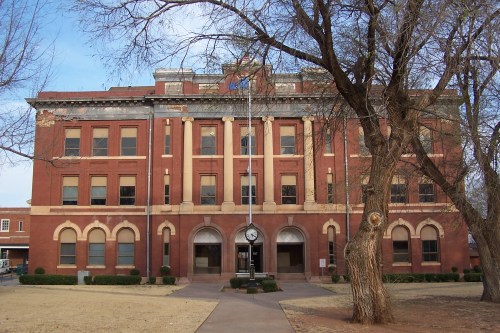 Historic brick building with a clock tower, surrounded by trees and grass, under a clear blue sky.