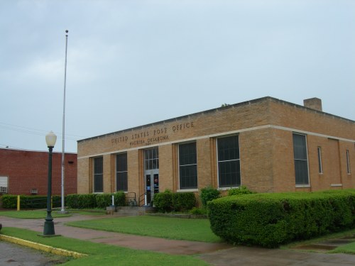 A brick building with large windows, labeled "United States Post Office," surrounded by green grass and shrubs.
