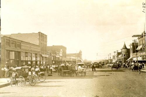 Historic street scene with horse-drawn carriages and people in early 20th-century attire, lined with buildings.