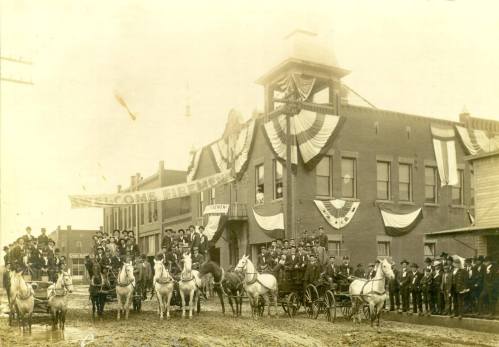 Historic black-and-white photo of a decorated building with a crowd, horses, and carriages during a celebration.