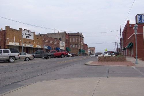 A quiet street scene featuring brick buildings, parked trucks, and storefronts with awnings in a small town.
