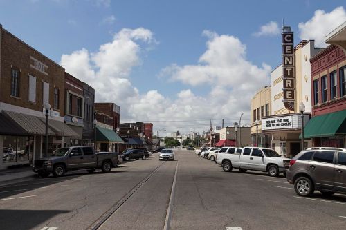 A small town street with shops, parked cars, and a theater marquee under a partly cloudy sky.