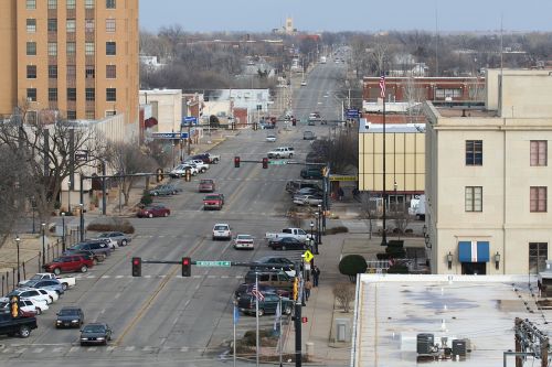 Aerial view of a city street with traffic lights, parked cars, and buildings lining both sides under a cloudy sky.