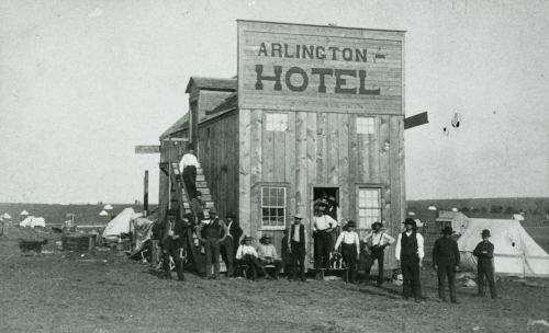 Historic photo of the Arlington Hotel with a group of people outside, surrounded by tents in a barren landscape.