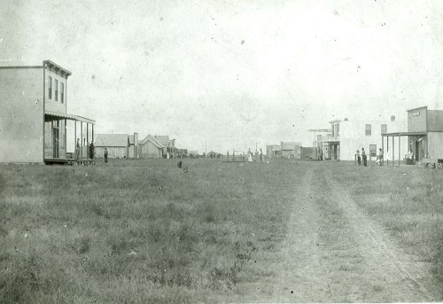 A historic black-and-white photo of a wide dirt street lined with wooden buildings and people in a grassy area.
