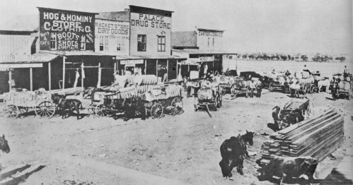 Historic black-and-white photo of a bustling frontier town with horse-drawn wagons and storefronts.