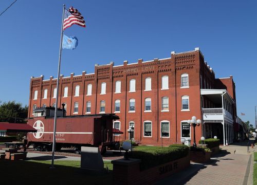 Historic red brick building with a flagpole and train car in front, set against a clear blue sky.