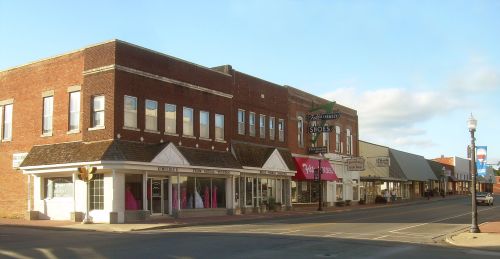 A quiet street scene featuring brick buildings with storefronts and a clear blue sky.