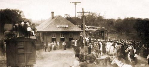 A historic train station bustling with people, horses, and wagons, capturing a lively scene from the past.