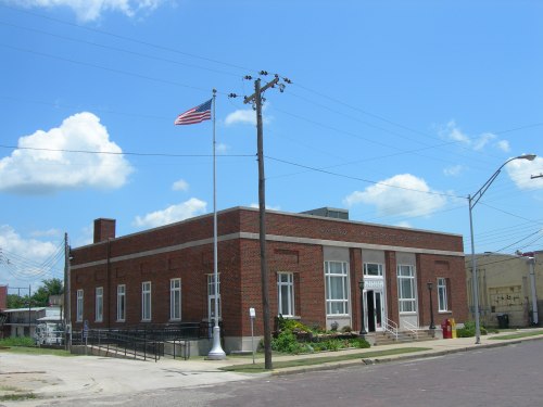 A brick building with large windows, an American flag, and a landscaped entrance under a blue sky with clouds.