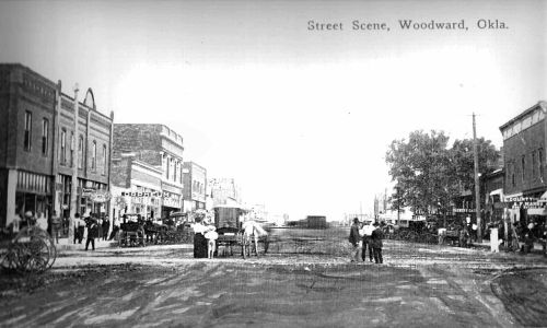 Historic street scene in Woodward, Oklahoma, featuring buildings, horse-drawn carriages, and people on a dirt road.