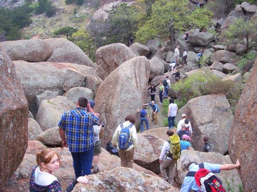 A group of people hiking through a rocky landscape with large boulders and greenery in the background.