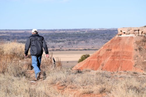 A person walks away on a grassy hill, with a red rock formation and expansive landscape in the background.