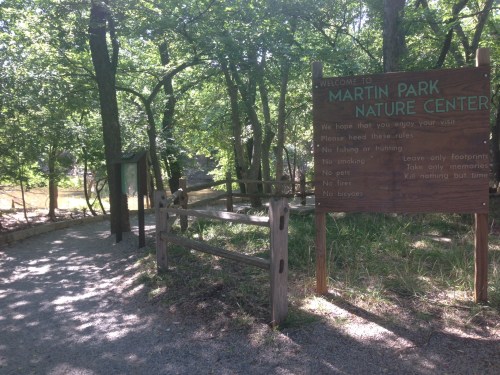 A path leads into a wooded area with a sign for Martin Park Nature Center, surrounded by trees and greenery.