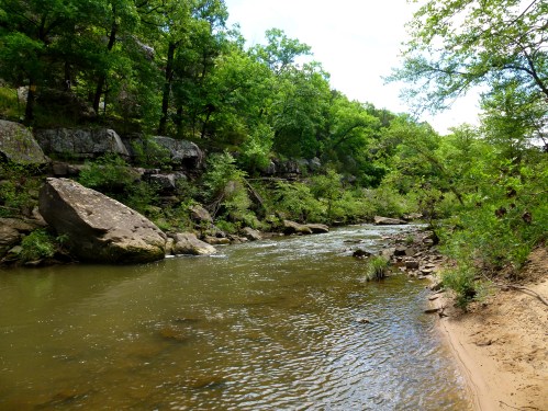 A serene river flows through a lush green landscape, surrounded by trees and rocky formations.