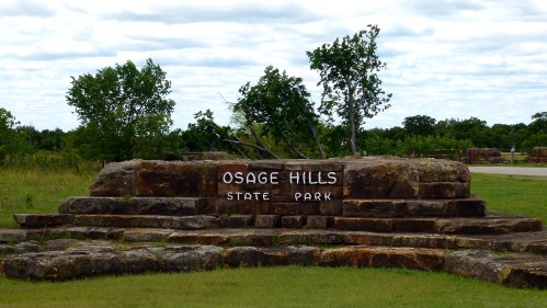 Sign for Osage Hills State Park on a stone structure, surrounded by grass and trees under a cloudy sky.
