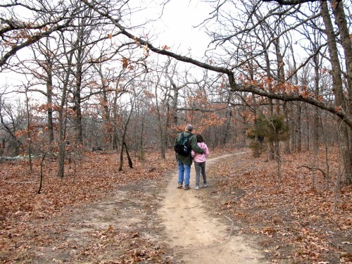A person and a child walk hand in hand along a dirt path in a wooded area with bare trees and fallen leaves.