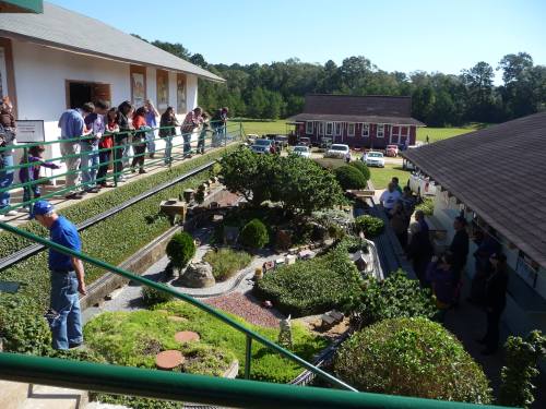 A view of a garden with people on a balcony above, surrounded by greenery and buildings in a sunny outdoor setting.