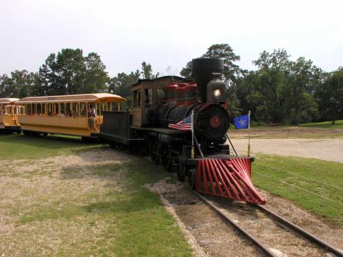 A vintage steam locomotive with a red front and yellow passenger cars on a gravel track surrounded by trees.