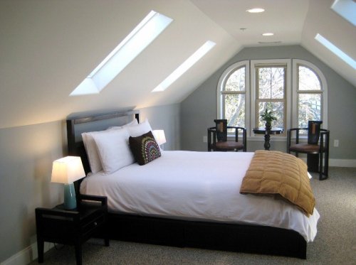 Cozy attic bedroom with a bed, two chairs, and large windows letting in natural light.
