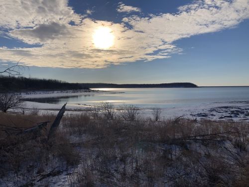 A serene winter landscape featuring a frozen shoreline, gentle waves, and a bright sun partially obscured by clouds.